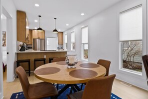A modern kitchen with stainless steel appliances, a dining area with a round table and chairs, and large windows with blinds. - The Gingerbread Flat | Storybook Charm in Downtown (Michigan City)