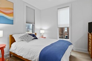 A bedroom with a bed, bedside lamps, a dresser, and a window with a view of a brick building. - The Gingerbread Flat | Storybook Charm in Downtown (Michigan City)