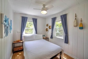 A bedroom with a bed, two windows with curtains, a ceiling fan, and a wall with decorative items. - Hamilton Hideaway | Walk to Beach & Fire Pit (Gary)