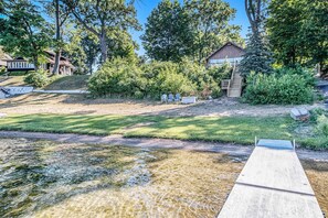 A riverside view with a wooden dock, grassy areas, and a building with a porch. - Hillside Cottage | 90 Ft Lake Frontage & Beach (Lawton)