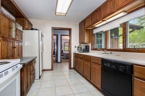 A kitchen with wooden cabinets, a refrigerator, a microwave, a dishwasher, and a window with a view of greenery. - Hillside Cottage | 90 Ft Lake Frontage & Beach (Lawton)