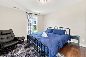 Ein Schlafzimmer mit einem Bett, einem Sessel, einem Nachttisch und einem Fenster mit Vorhängen.