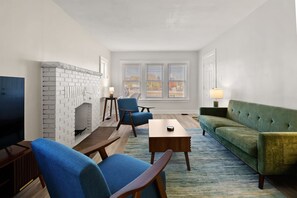 A living room with a brick fireplace, a television, a blue armchair, a green sofa, and a wooden coffee table.