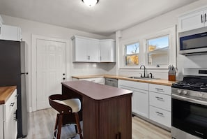 A modern kitchen with white cabinetry, a wooden bar stool, a stainless steel stove, and a window with autumn leaves. - Main Street Retreat | Mid-century charm steps from the lake and arts district (Michigan City)