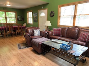 A cozy living room with a brown leather sofa, a matching armchair, a coffee table, and a dining area with a red and black checkered tablecloth. - Mt. Baldy Cabin | Game Room, Fire Pit & The Dunes (Michigan City)