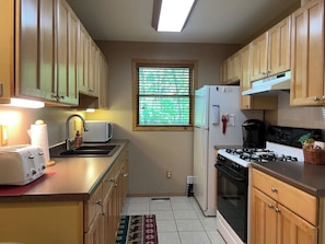 A compact kitchen with wooden cabinets, a gas stove, a refrigerator, and a window with blinds. - Mt. Baldy Cabin | Game Room, Fire Pit & The Dunes (Michigan City)