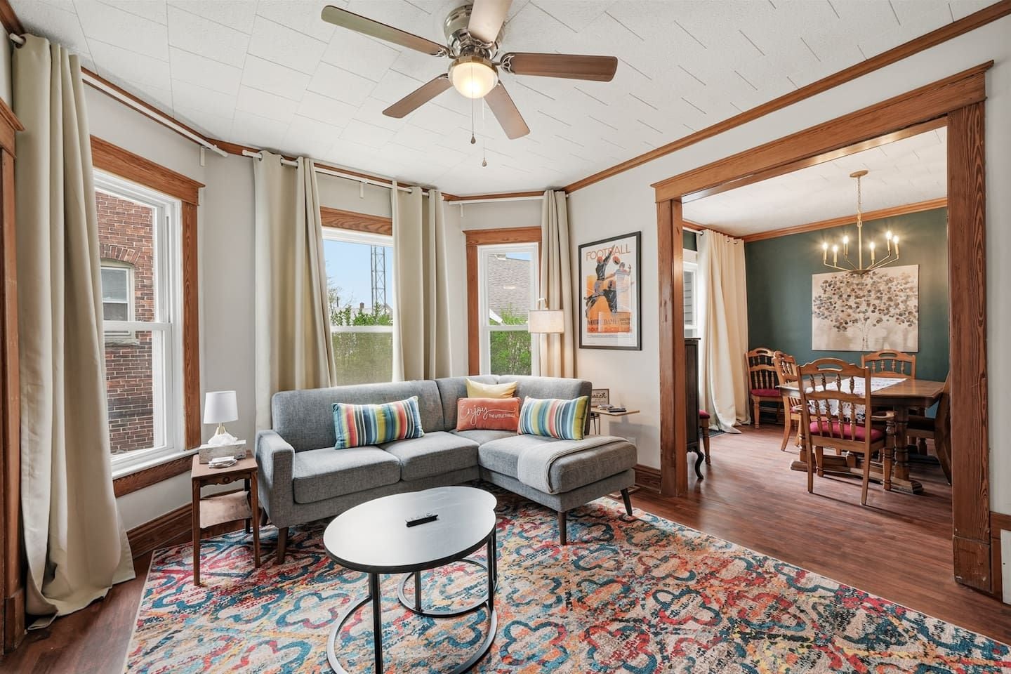 A living room with a gray sectional sofa, a round coffee table, and a colorful rug. There is a dining area with wooden chairs and a table.
