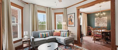A living room with a gray sectional sofa, a round coffee table, and a colorful rug. There is a dining area with wooden chairs and a table.