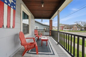 A covered porch with red Adirondack chairs, a small table, and a view of a residential street. - Pine Street Flat | Steps to Everything (Michigan City)