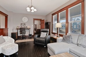 Un salon avec un fauteuil en cuir noir, un canapé gris et une table basse en bois. On y trouve également un bureau avec une chaise et une lampe.