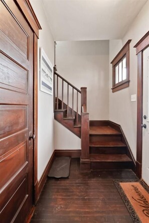 A wooden staircase with a handrail leading to a landing, a door on the left, and a window on the right. - The Porch | Craftsman Charm & Steps to Downtown (Michigan City)