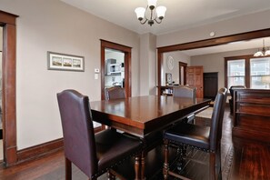 A dining room with a large wooden table and leather chairs, a chandelier, and a view into the kitchen. - The Porch | Craftsman Charm & Steps to Downtown (Michigan City)