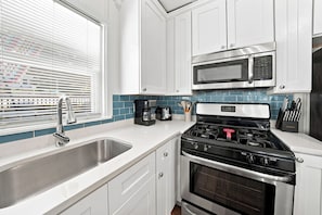 A modern kitchen with white cabinets, a stainless steel sink, a microwave, a gas stove, and a coffee maker. - Sheridan Beach Bungalow (Michigan City)
