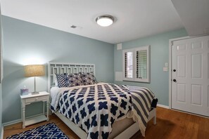 A bedroom with a bed, bedside table, lamp, and a window with blinds. - The Tree House | Lake Views & Beach a Block Away (Sawyer)