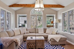 A modern living room with a sectional sofa, a hanging light fixture, and large windows offering a view of the outdoors. - The Tree House | Lake Views & Beach a Block Away (Sawyer)