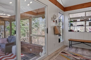 A room with a large sliding glass door leading to a deck, a bench, a shelf with decorative items, and a patterned rug. - The Tree House | Lake Views & Beach a Block Away (Sawyer)