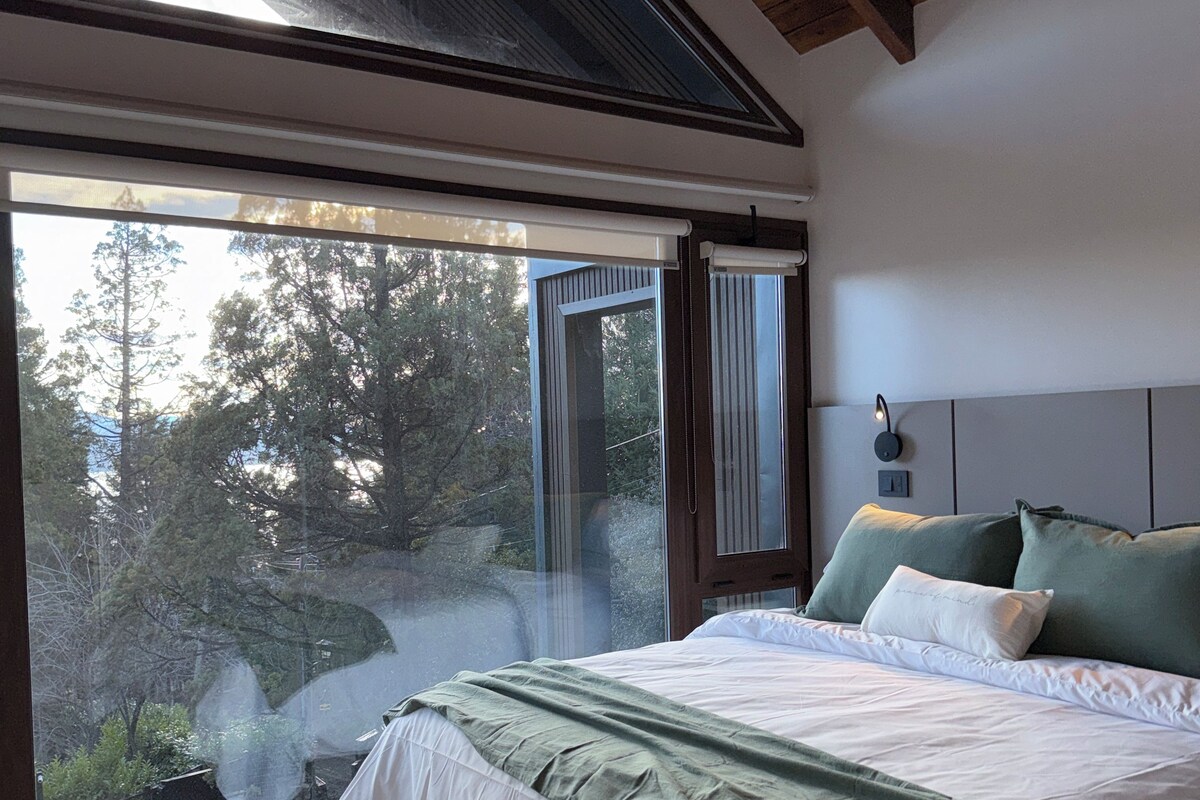 A bedroom with a large window, a bed with green bedding, and a hanging light fixture.