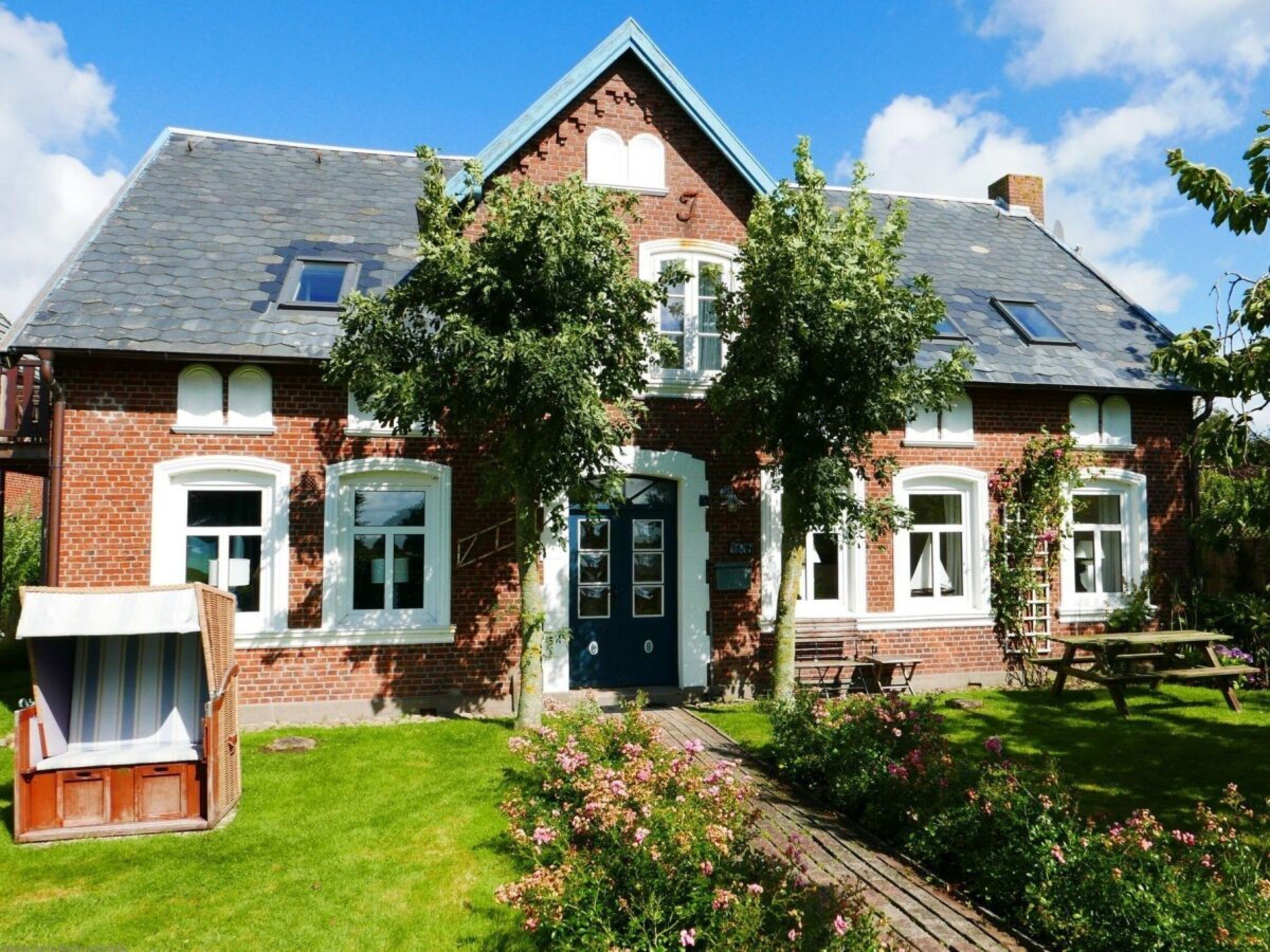 Une maison à deux étages, construite en briques, avec un toit à pignon, une porte d’entrée et un petit porche agrémenté d’un banc.