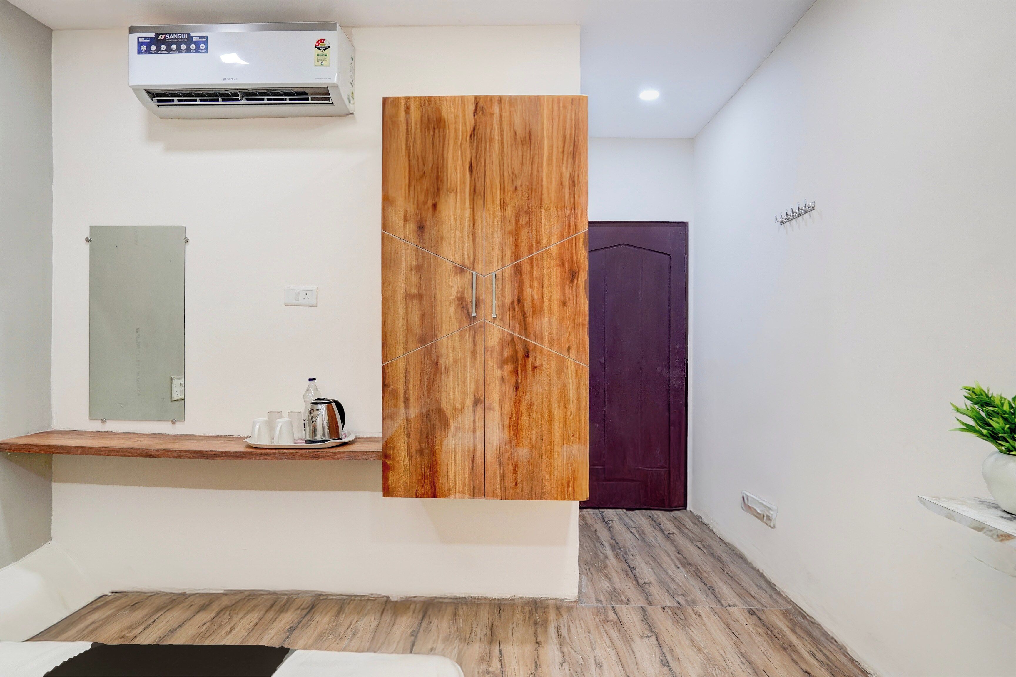 A hotel room with a wooden wardrobe, a small table with a kettle and cups, a mirror, and a purple door.