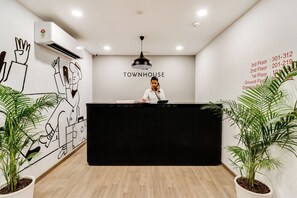 A reception desk with a person behind it, a wall with a 'TOWNHOUSE' sign, and a list of phone numbers on the right. - Townhouse Gandhi Hall (Indore)