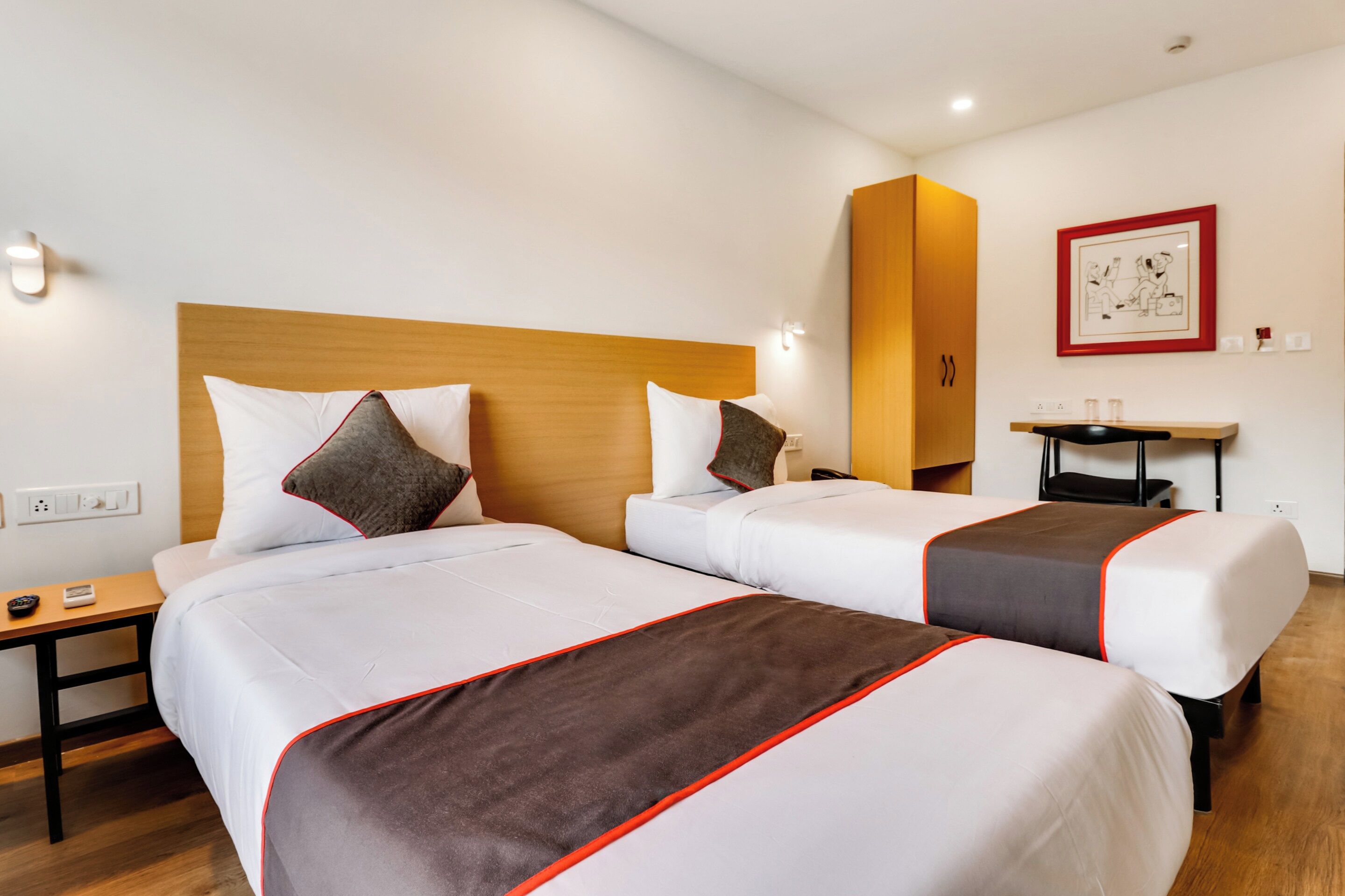 A hotel room with two beds, a wooden headboard, bedside tables, a desk, a chair, and a framed picture on the wall.