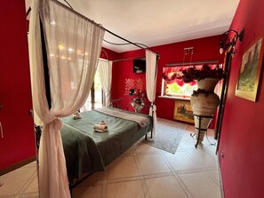 A room with a bed, a window with red curtains, a vase with flowers, and a painting on the wall.