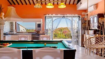 A pool table in a room with a view of the outdoors, a kitchen area, and a dining space with a view.