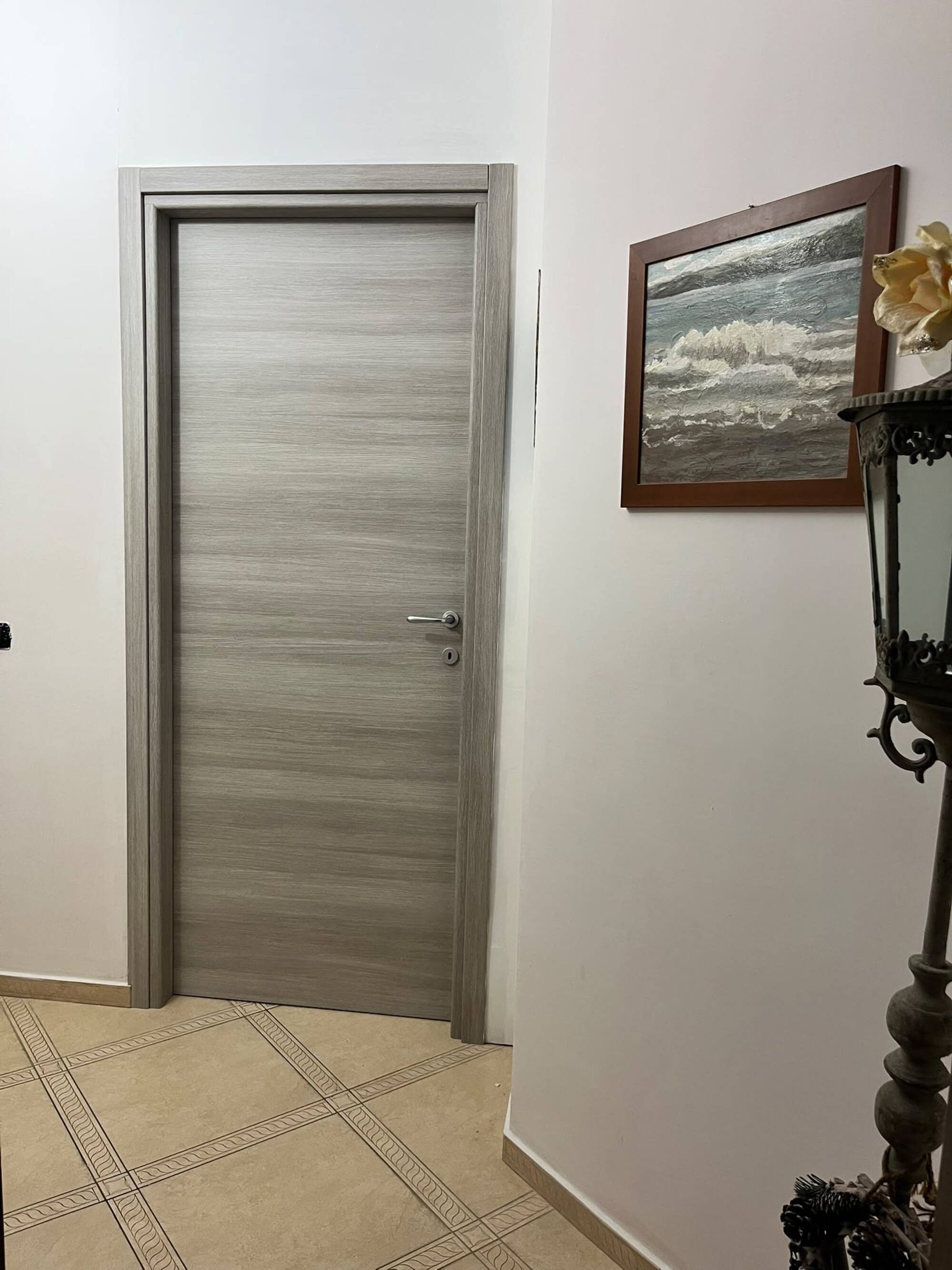 Una puerta cerrada con superficie texturizada, un cuadro enmarco con una vista marina en la pared y una lámpara decorativa con un arreglo floral.
