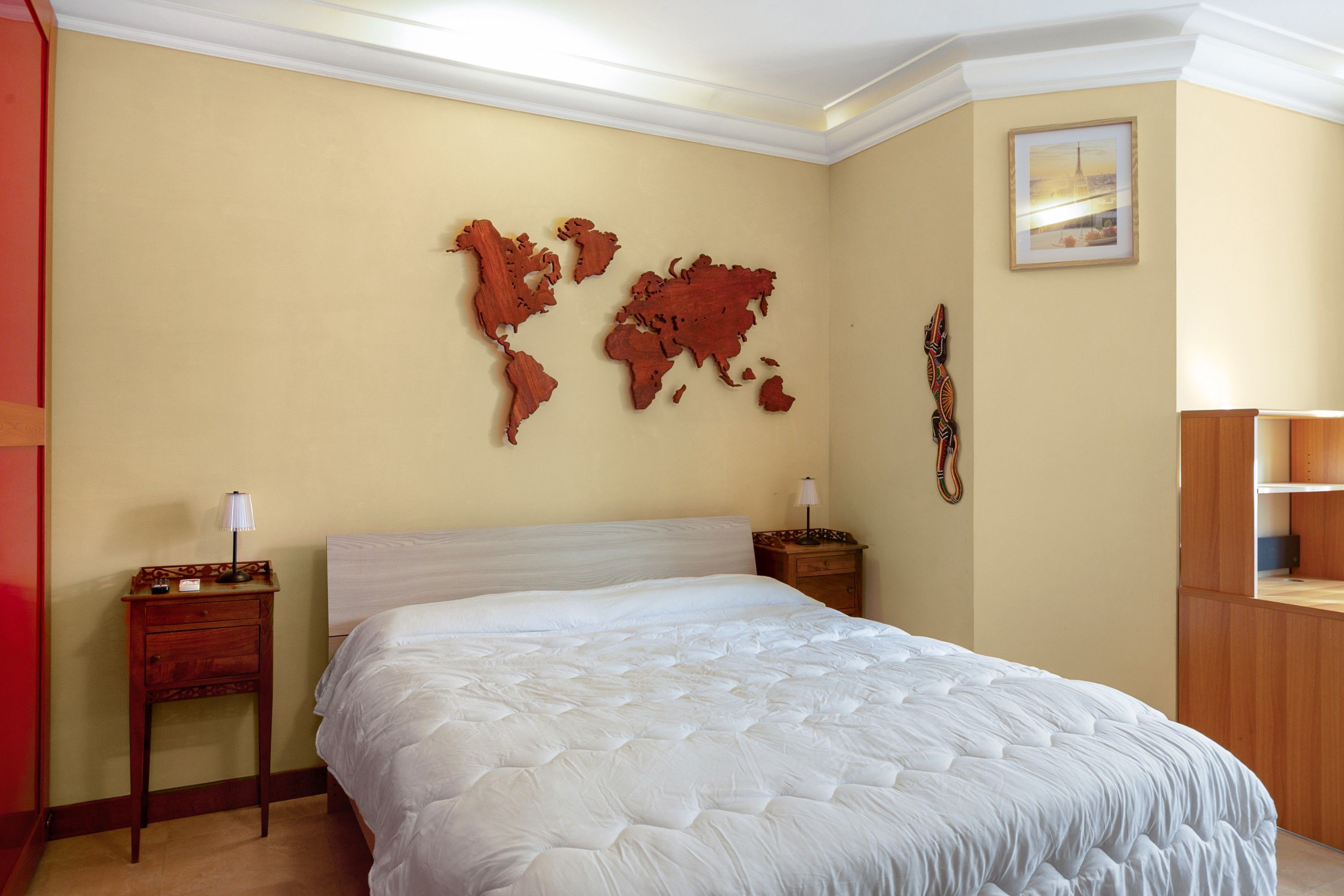 Una habitación de hotel con una cama grande, dos mesitas de noche, una obra de arte con un mapa mundial, un cuadro enmarcado y un armario de madera.