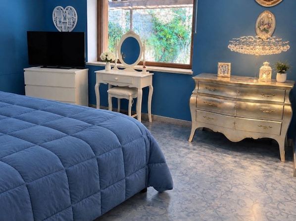 A bedroom with a blue bed, a white dresser, a vanity with a mirror, and a television on a stand.
