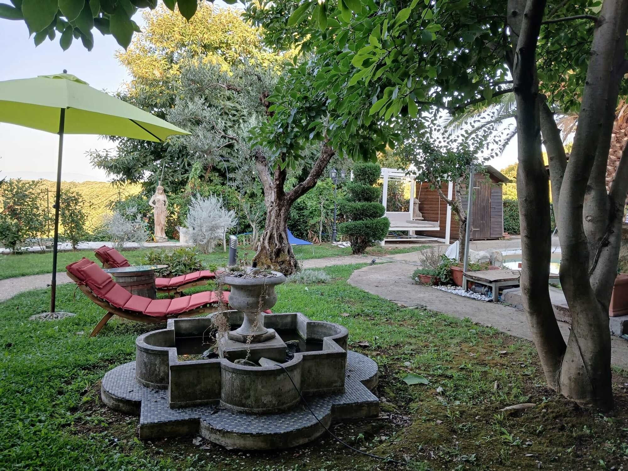 Ein Garten mit einem runden Steinfountain, roten Liegestühlen und einem grünen Sonnenschirm.