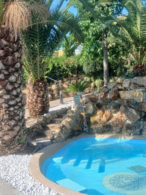 Un área de piscina con pared de piedra, palmeras y una cascada.