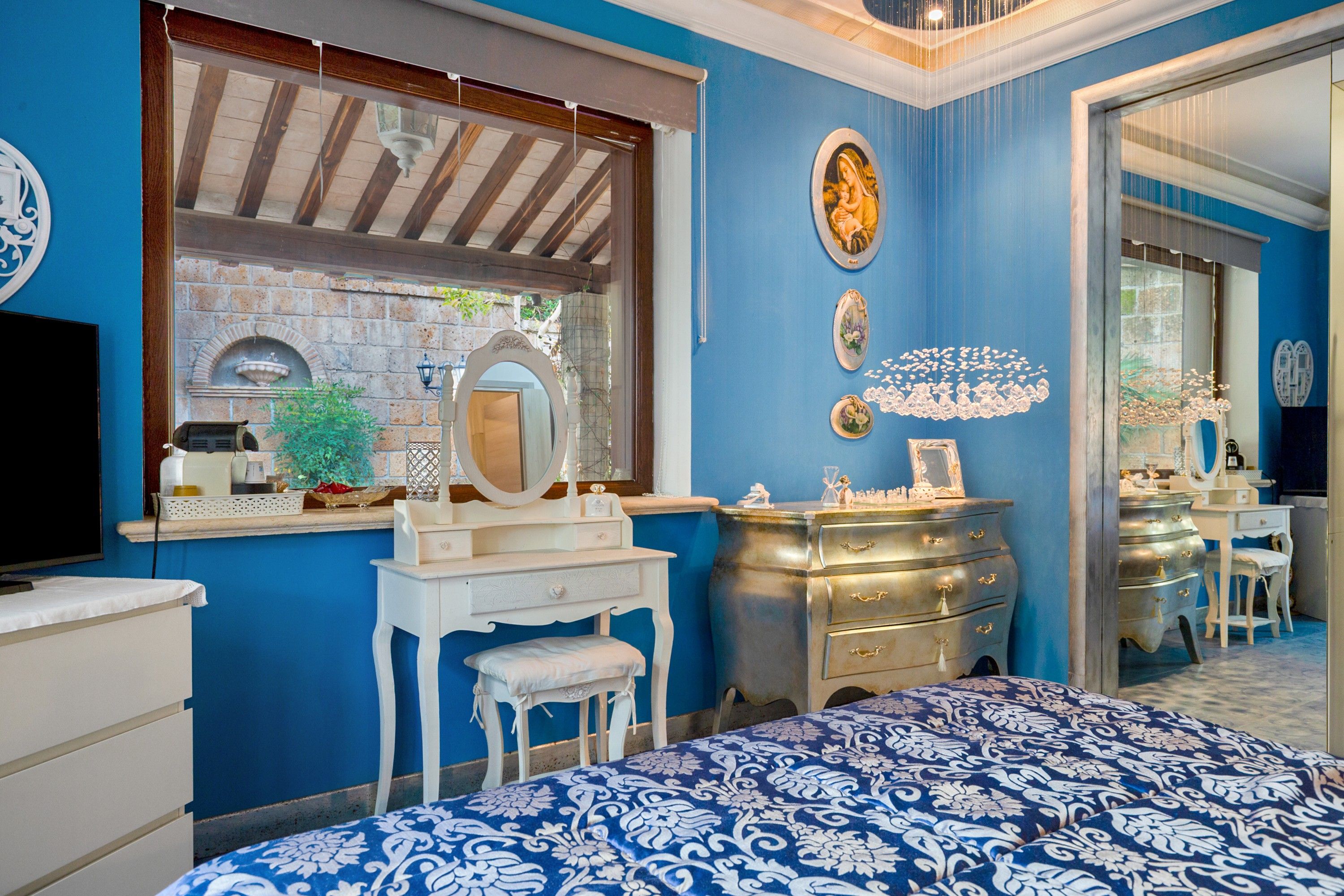 Une chambre à coucher comprenant un lit, une commode, une coiffeuse avec un miroir et une fenêtre donnant sur un mur de briques et de la verdure.
