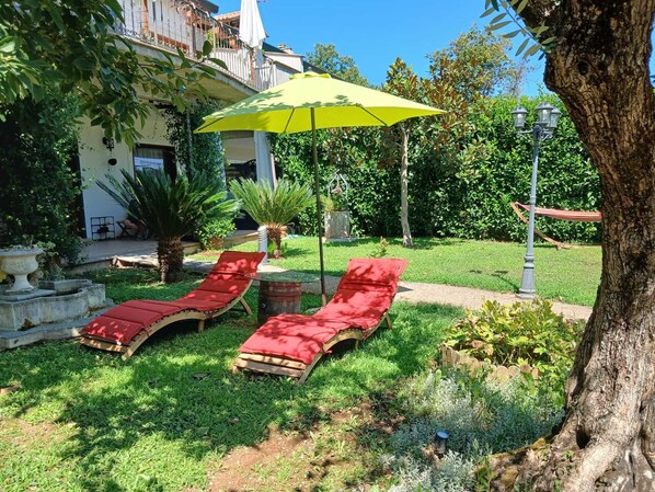 Un jardín con dos sillones rojos bajo un paraguas amarillo, rodeado de vegetación y un edificio al fondo.