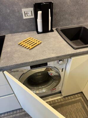Ein kleiner Waschbereich mit Waschmaschine, Spüle und einer Arbeitsplatte, versehen mit Papierhandtuchspender und Holz-Wäscheklammern.