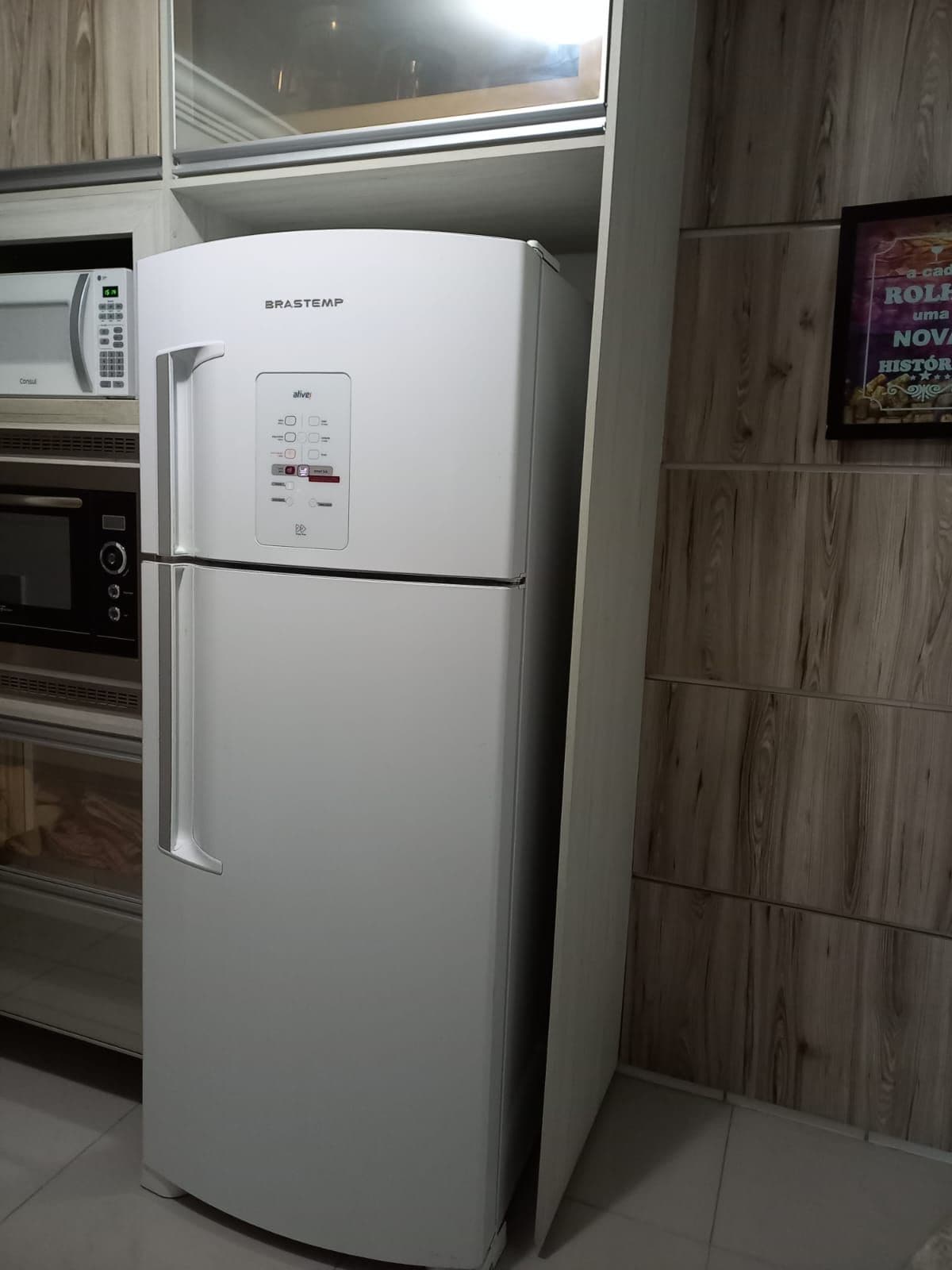 A white refrigerator with a digital control panel, a microwave above it, and a wooden wall panel.