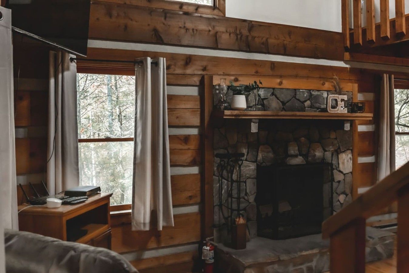 A cabin interior with a stone fireplace, wooden beams, a window with curtains, and a desk with a computer.