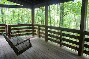Een houten terras met een schommel en een bankje, omgeven door een houten balustrade, met uitzicht op een bebost gebied.