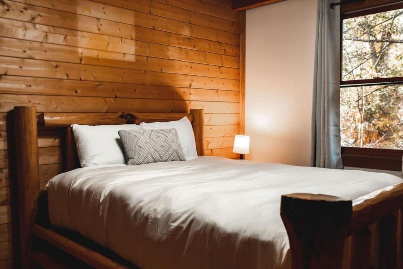 A wooden-paneled room with a bed, a nightstand, and a window with a view of trees.