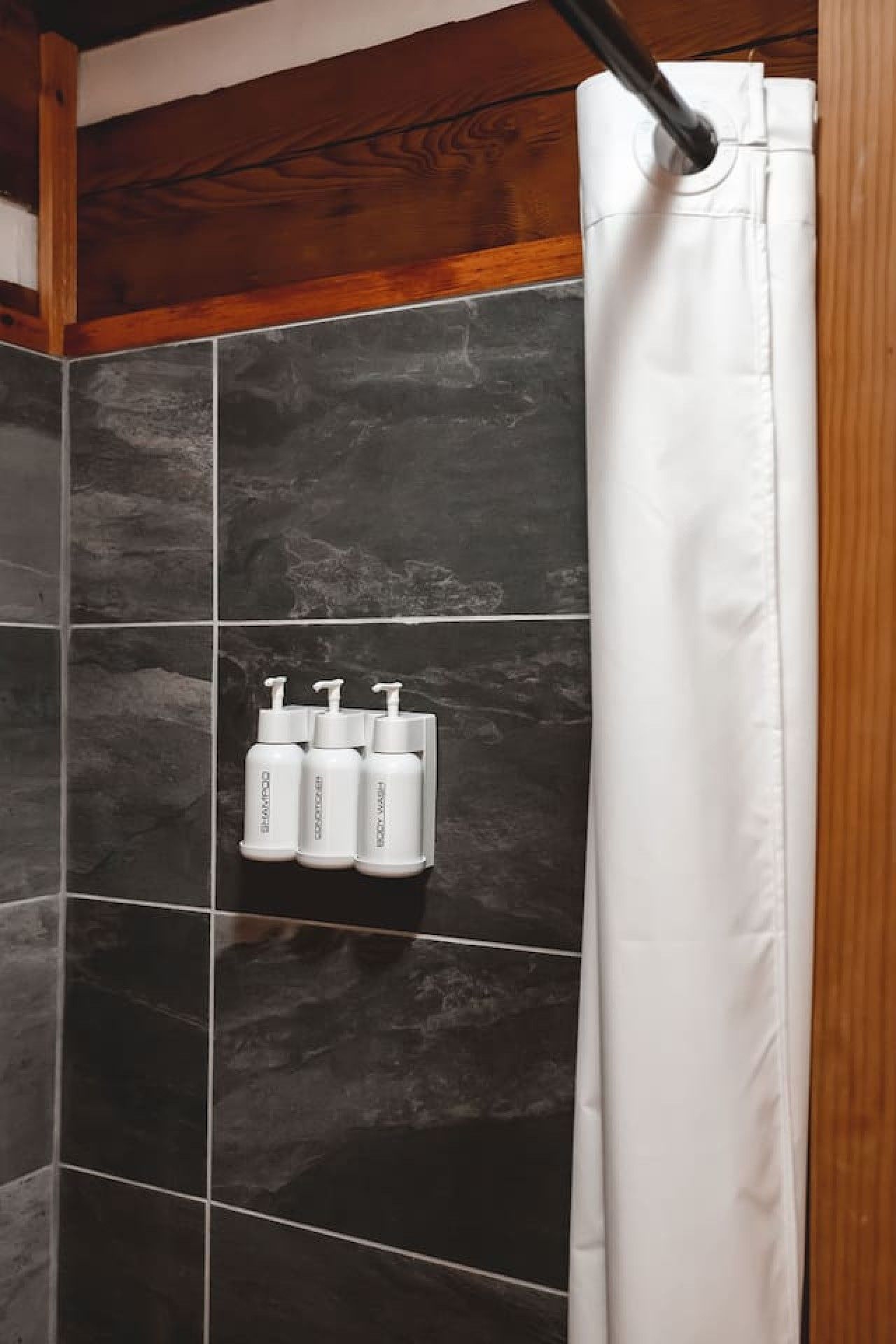 Un baño con baldosas oscuras, techo de madera y cortina de ducha blanca. Hay tres botellas blancas en la pared, probablemente con artículos de aseo.