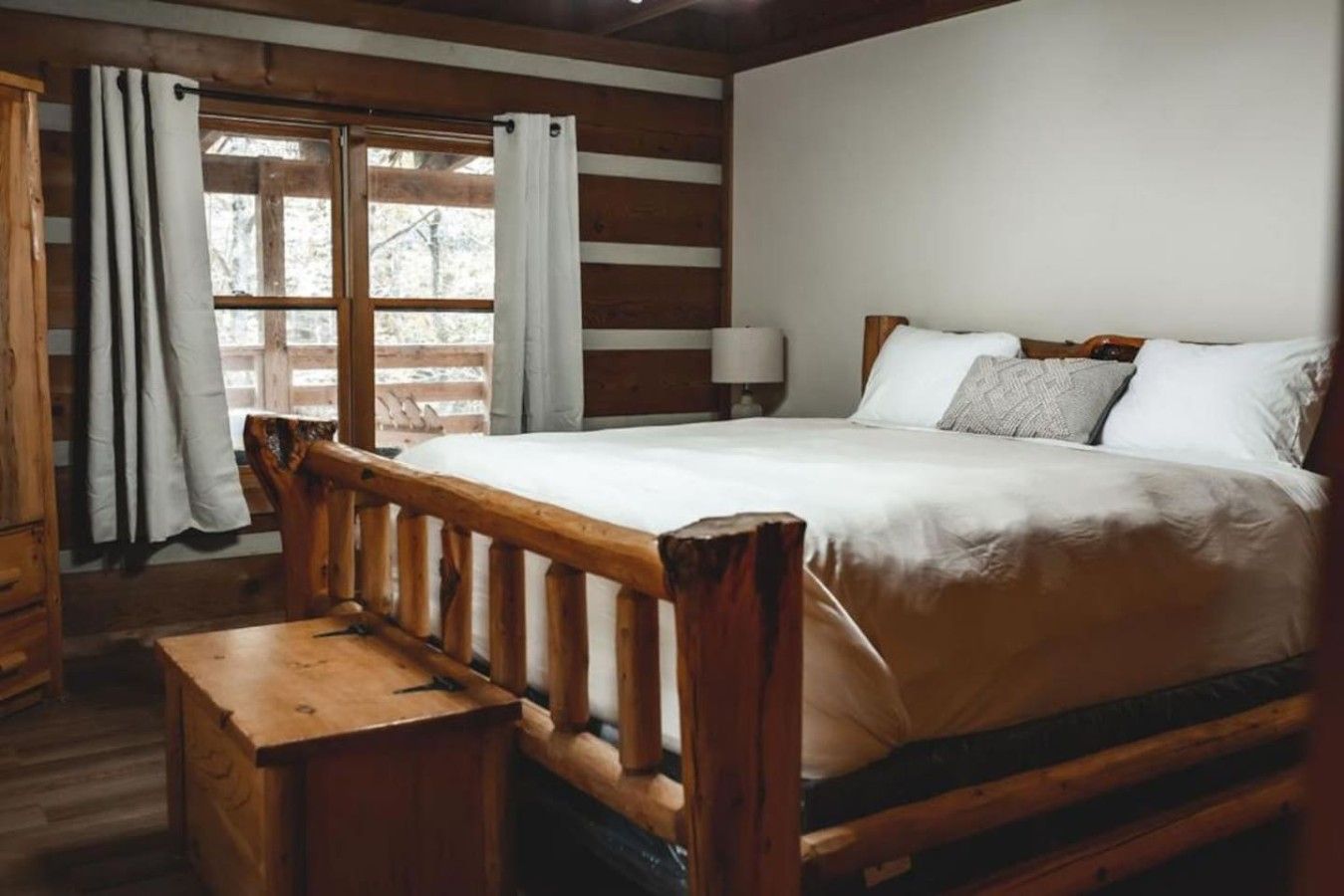 Een bed met houten frame, een wit matras en kussens, een houten bankje, een nachtkastje en een raam met gordijnen.