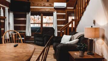 L’intérieur d’un chalet accueillant, avec une table à manger en bois, un canapé, un fauteuil, une lampe et un escalier.