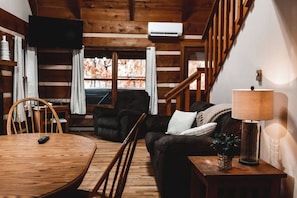L’intérieur d’un chalet accueillant, avec une table à manger en bois, un canapé, un fauteuil, une lampe et un escalier.