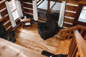L'intérieur d'une cabane avec un plancher en bois, un fauteuil noir, une petite table avec une lampe et une fenêtre avec des rideaux.