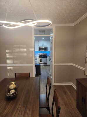 Un comedor con mesa y sillas de madera, un cuenco decorativo pequeño y vista a una cocina con armarios y electrodomésticos.