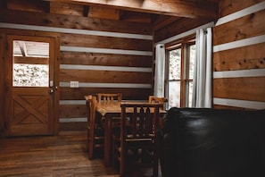 Een houten chaletinterieur met een eettafel en stoelen, een zwarte leren bank en een raam met gordijnen.