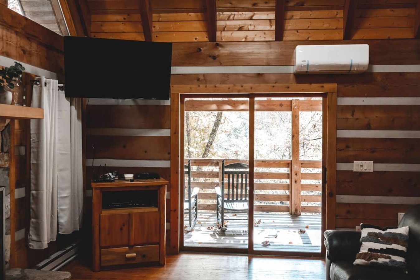 A cabin interior with a wooden floor, a TV mounted on the wall, a sliding glass door leading outside, and a black leather sofa.