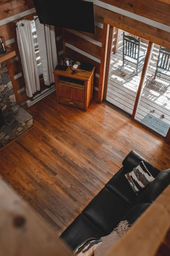 Een woonkamer met houten vloeren, een zwarte bank, een houten kast en een aan de muur bevestigde televisie.