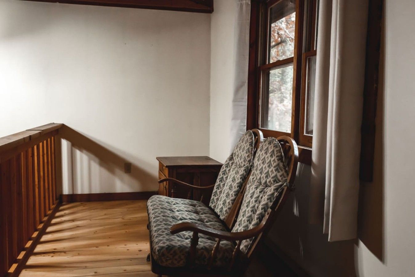 Une pièce d’angle avec un plancher en bois, une chaise, une petite table et une fenêtre avec des rideaux.