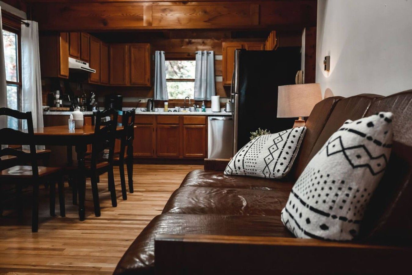 Een gezellige woonkamer met een leren bank, gestreepte kussens, een eethoek met houten stoelen, en een keuken met houten kastjes en een koelkast.